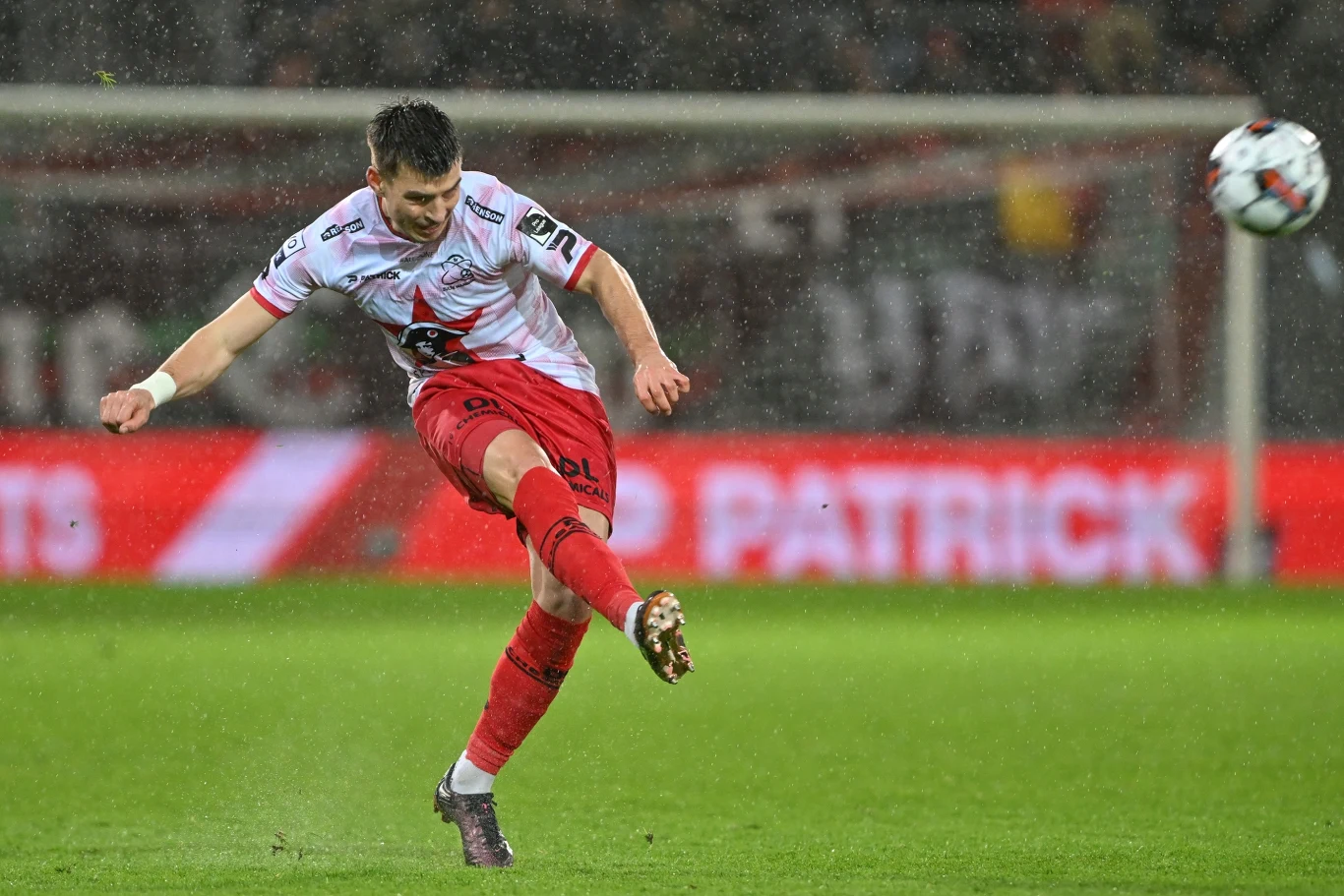 Karol Fila Piłkarz w czerwono-białym stroju wykonuje silny strzał w trakcie meczu piłki nożnej, w tle widoczny rozmyty stadion i reklamy.