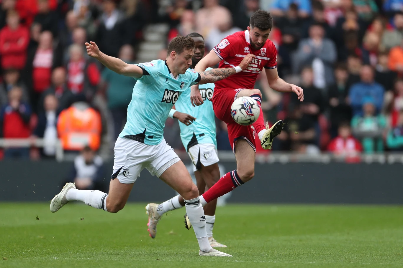 Dwóch piłkarzy rywalizujących o piłkę podczas meczu na stadionie, otoczeni przez innych zawodników i kibiców na trybunach, akcja dynamiczna, po lewej zawodnik w jasnym stroju, po prawej w czerwonym.