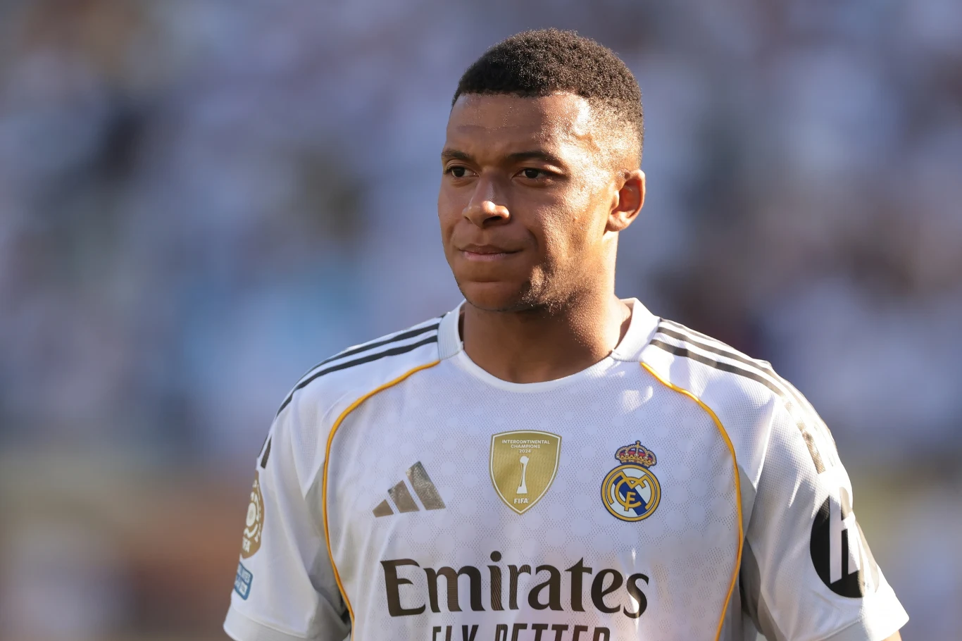 Kylian Mbappe Piłkarz w białym stroju Realu Madryt z logo sponsora i złotymi akcentami, skupiony, stoi na tle rozmytego stadionu w świetle dziennym.
