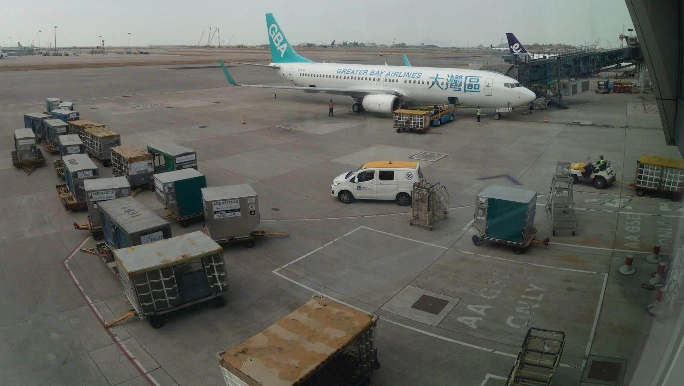 Tanie linie lotnicze Greater Bay Airlines musiały ograniczyć kursy na trasach do Japonii, a niektóre, jak te do Tokushimy, nawet całkowicie zawiesić. Samolot pasażerski Greater Bay Airlines zaparkowany przy terminalu lotniska, obok samolotu znajduje się pojazd obsługi naziemnej, w tle rozmieszczone są kontenery bagażowe i sprzęt lotniskowy, wszystkie elementy wskazują na standardowy ruch lotniczy.