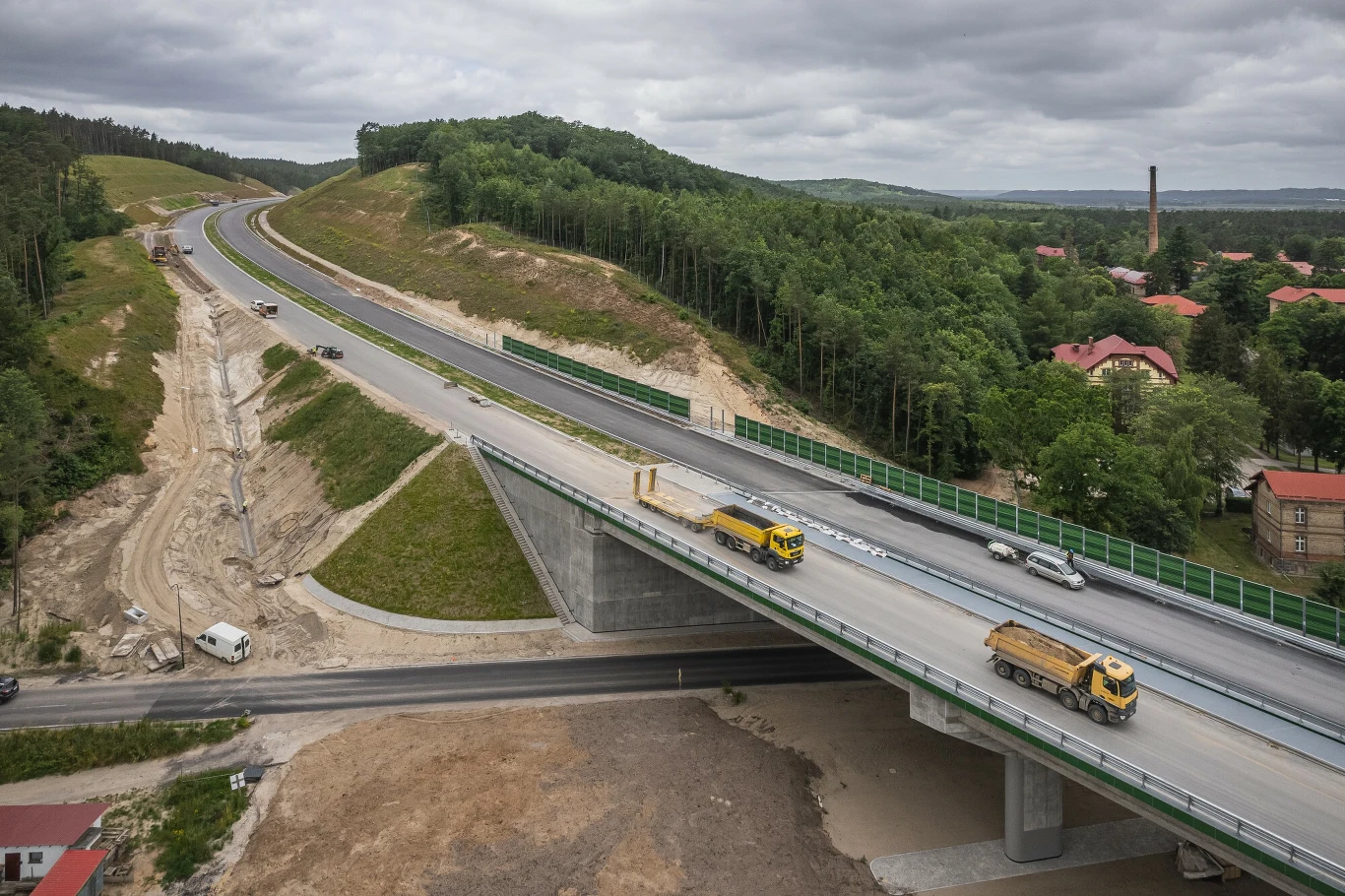 Budowa trasy S6 Nowoczesny most drogowy z przejeżdżającymi ciężarówkami i samochodami, prowadzący przez tereny leśne, w tle widać zielone wzgórza, domy z czerwonymi dachami oraz industrialny komin.