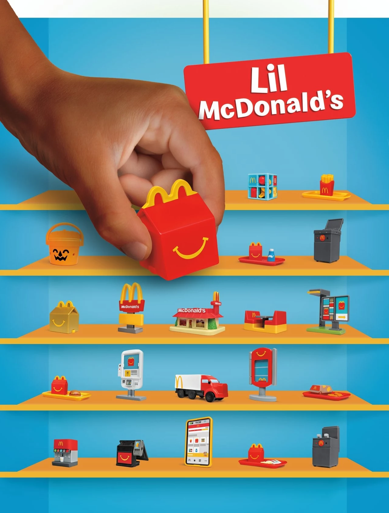Ręka sięgająca po małe opakowanie Happy Meal w otoczeniu miniaturowych zabawek i akcesoriów związanych z restauracją McDonald's, takich jak restauracja, samochód dostawczy, kasa czy zestawy jedzenia na półkach.