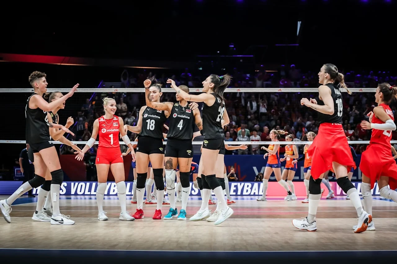Zawodniczki dwóch drużyn siatkarskich, ubrane w stroje sportowe, znajdują się na boisku hali podczas meczu, część z nich wyraża radość lub gratulacje wobec siebie nawzajem, w tle widoczna publiczność i siatka.