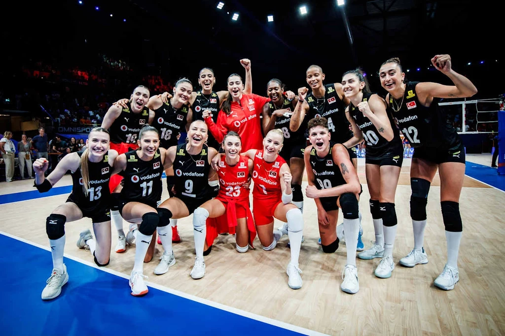 Grupa zawodniczek siatkarskich na hali sportowej, ubrane w stroje meczowe w kolorach czarnym i czerwonym, stoją razem i cieszą się po meczu, unosząc ręce i okazując radość, w tle boisko oraz trybuny.