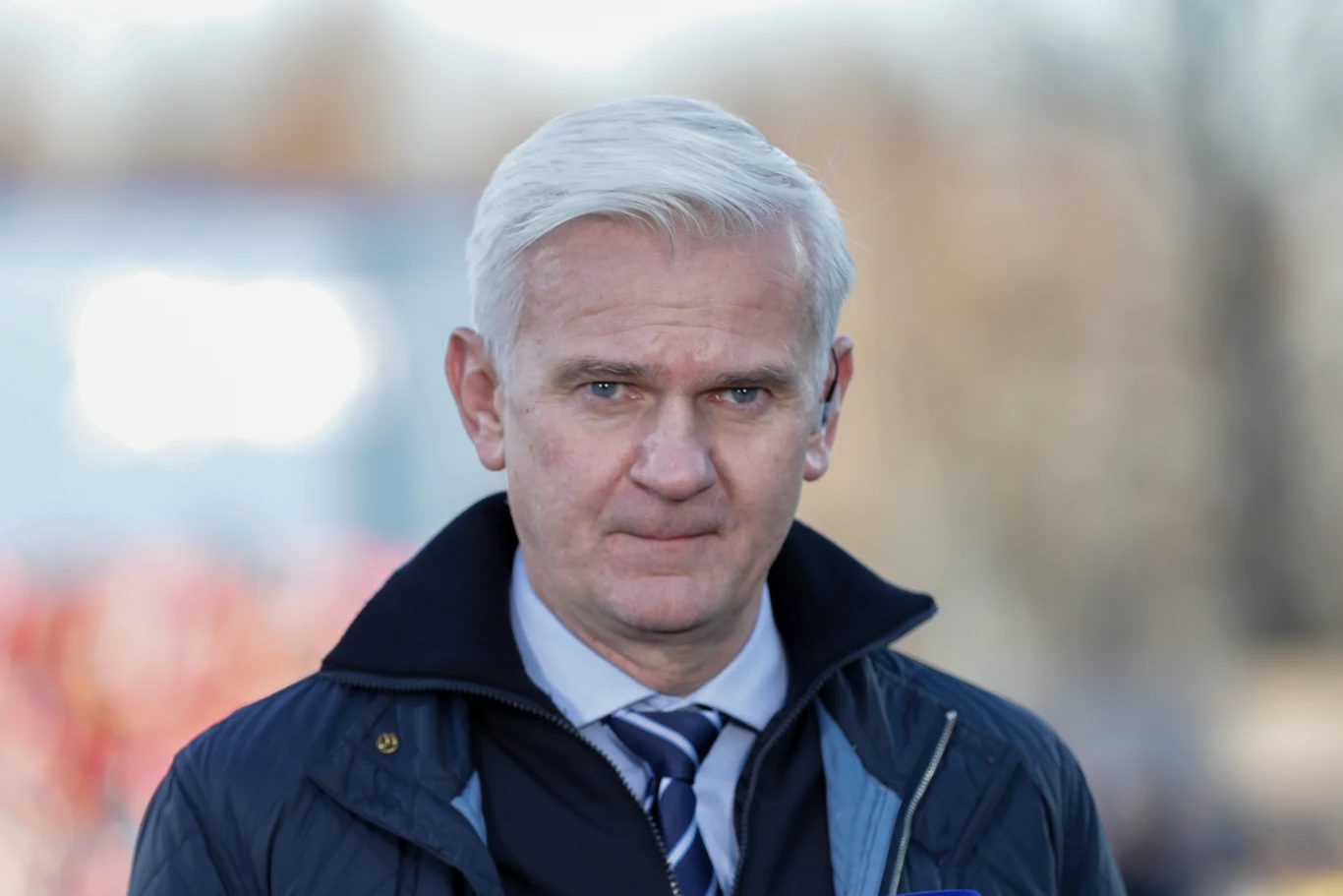 Mężczyzna z siwymi włosami ubrany w granatowy płaszcz oraz krawat, patrzy poważnie w stronę obiektywu, rozmyte tło w jasnych kolorach.