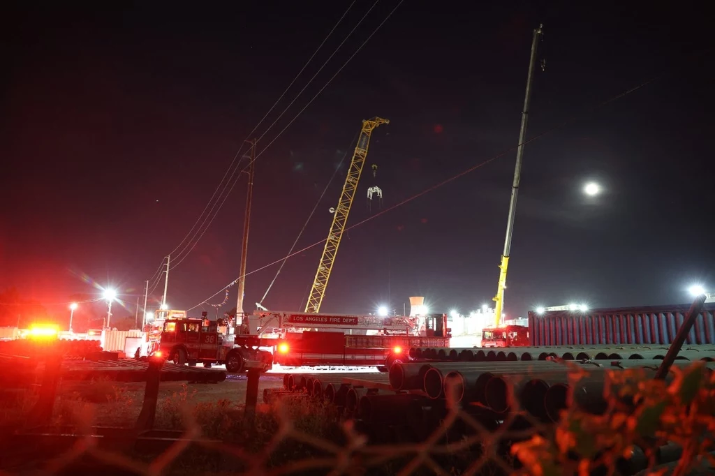 Zespoły poszukiwawczo-ratownicze oraz specjalny dźwig na miejscu akcji w Los Angeles Rozświetlony plac budowy nocą, widoczne dwa duże dźwigi pracujące przy przenoszeniu materiałów, kilka wozów strażackich oraz rury ułożone po prawej stronie kadru, w tle jasne światła i księżyc na niebie.