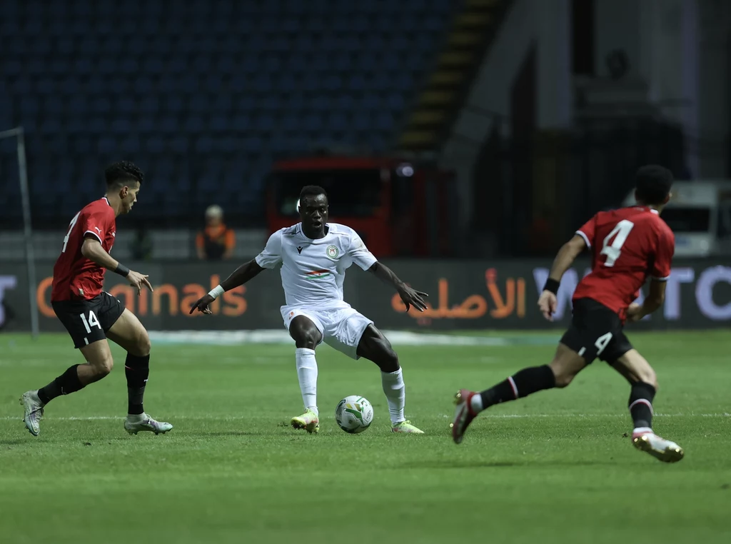 Daniel Sosah (w środku) Trzech piłkarzy w trakcie meczu piłki nożnej na stadionie, zawodnik w białym stroju drybluje piłkę między dwoma zawodnikami w czerwono-czarnych strojach, dobrze oświetlone boisko, trybuny w tle.