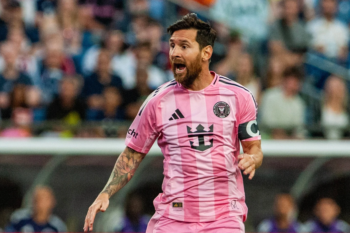 Leo Messi Piłkarz w różowej koszulce z opaską kapitana, dynamicznie gestykulujący podczas meczu piłkarskiego, w tle trybuny wypełnione kibicami.