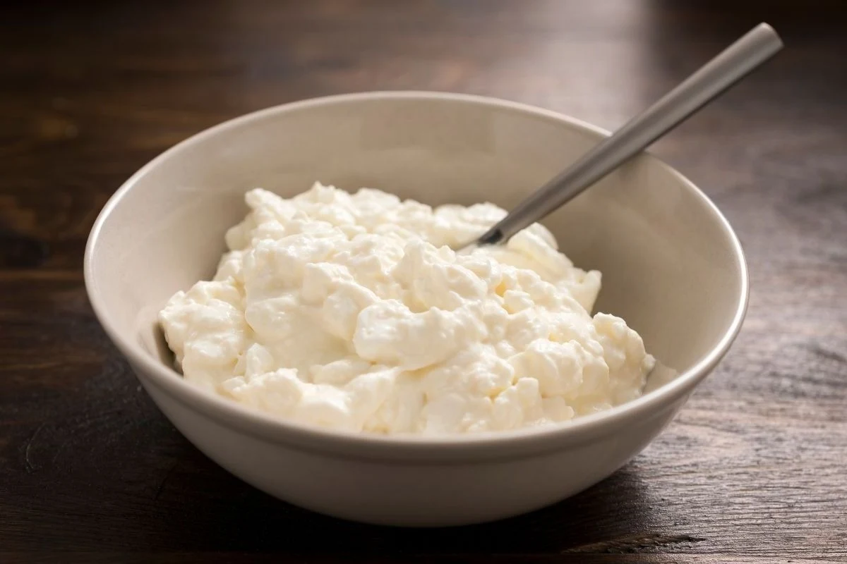 Ciasto z serka wiejskiego to zdrowy wybór na święta Biała miska wypełniona twarogiem ziarnistym z metalową łyżką, ustawiona na drewnianym blacie stwarza wrażenie świeżości i prostoty.