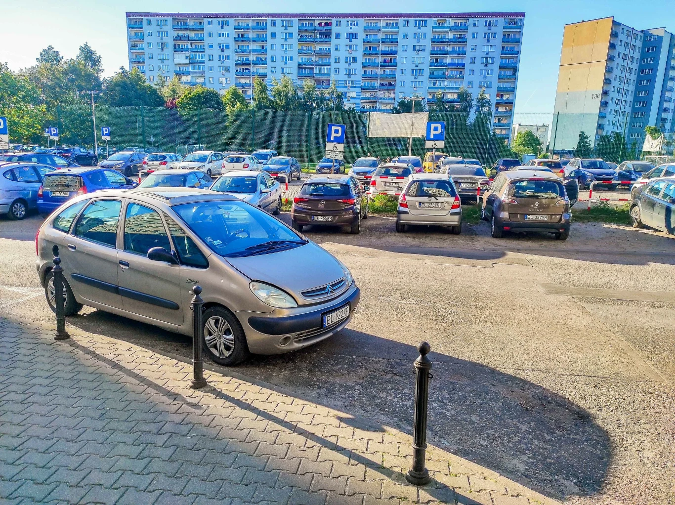 Koniec obowiązkowych parkingów przy nowych blokach. Deweloperzy zyskają Szare auto osobowe zaparkowane na ulubionym miejscu na skraju chodnika, liczne inne pojazdy stojące na parkingu miejskim, w tle wielopiętrowe bloki mieszkalne oraz oznakowanie parkingu.