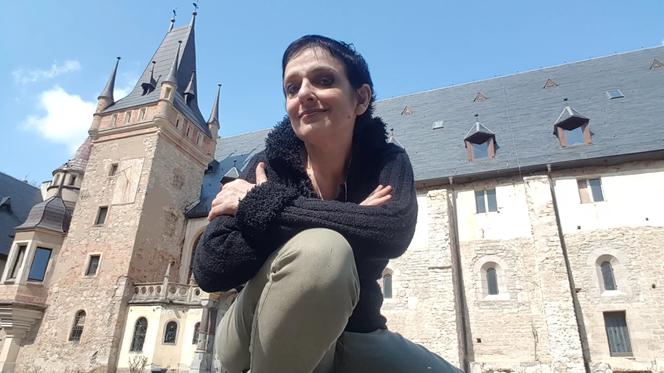 Kobieta o krótkich ciemnych włosach, ubrana w czarną bluzę i jasnozielone spodnie, siedzi z założonymi rękami przed zabytkowym budynkiem o kamiennych murach i wieżach na tle błękitnego nieba.
