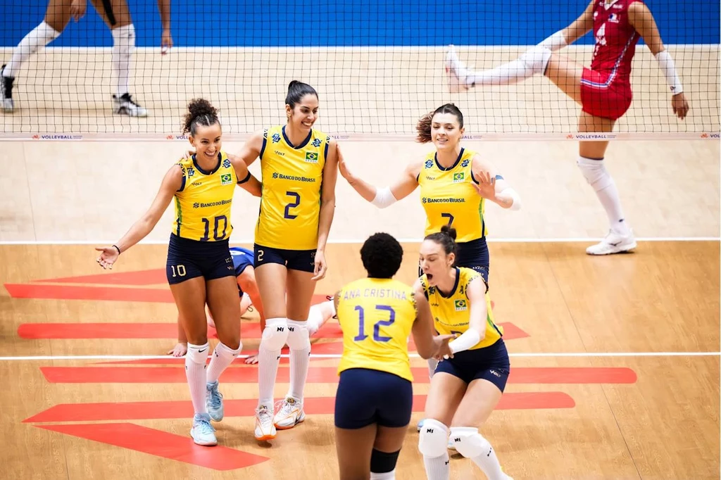 Siatkarki reprezentacji Brazylii w meczu Ligi Narodów z Francją Grupa zawodniczek ubranych w żółto-niebieskie stroje siatkarskie świętuje zdobyty punkt na parkiecie, jedna z zawodniczek drużyny przeciwnej w czerwonym stroju opuszcza boisko w tle za siatką.