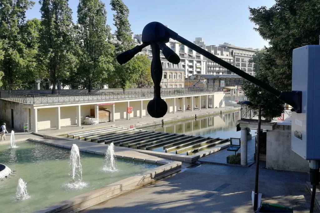 Miejski kanał wodny z rzędami wodotrysków otoczony drzewami i budynkami, na pierwszym planie widoczne nowoczesne urządzenie pomiarowe zamontowane na wysięgniku.
