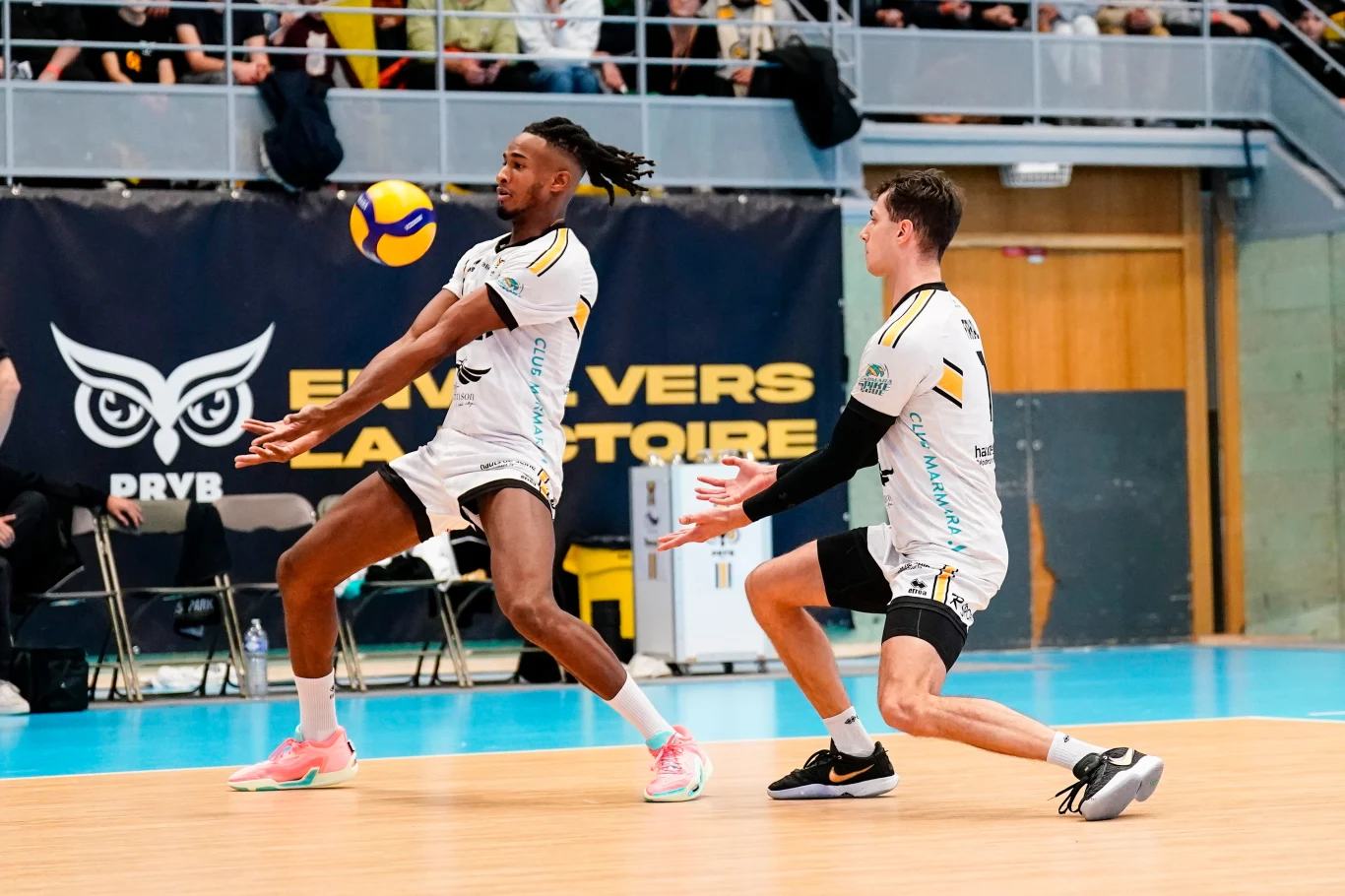 Samuel Jeanlys (z lewej) zagra w PlusLidze Dwóch siatkarzy w trakcie odbioru piłki na hali sportowej, skupieni na akcji, w tle widoczna publiczność oraz ławka rezerwowych.