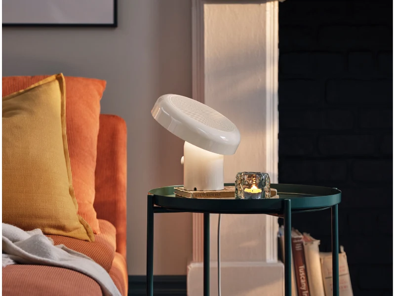 Głośnik Bluetooth z lampką Ikea Blomprakt. Pomarańczowa sofa z żółtą poduszką, mały okrągły stolik z zielonym blatem, na którym stoi biała lampa o nowoczesnym kształcie oraz przezroczysty świecznik z zapaloną świeczką. W tle jasna ściana i fragment czarnego kominka.
