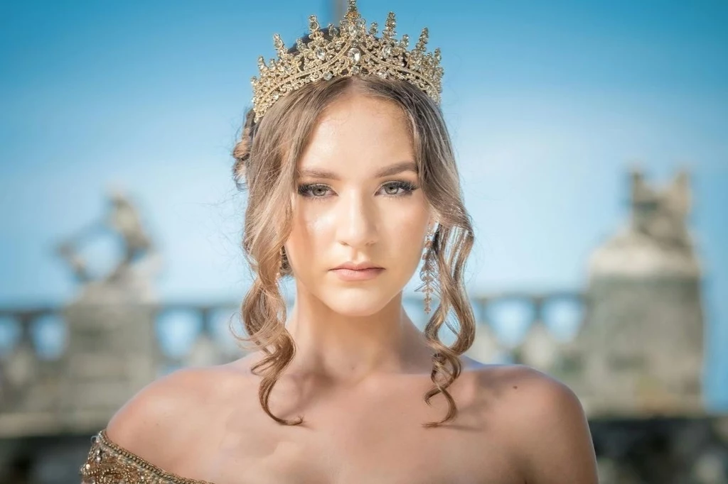 Imiona inspirowane baśniami szczególnie chętnie nadaje się dziewczynkom Młoda kobieta w ozdobnej złotej koronie na głowie, z włosami upiętymi w delikatne loki, patrząca poważnym wzrokiem w obiektyw na tle zamazanej architektury i nieba.