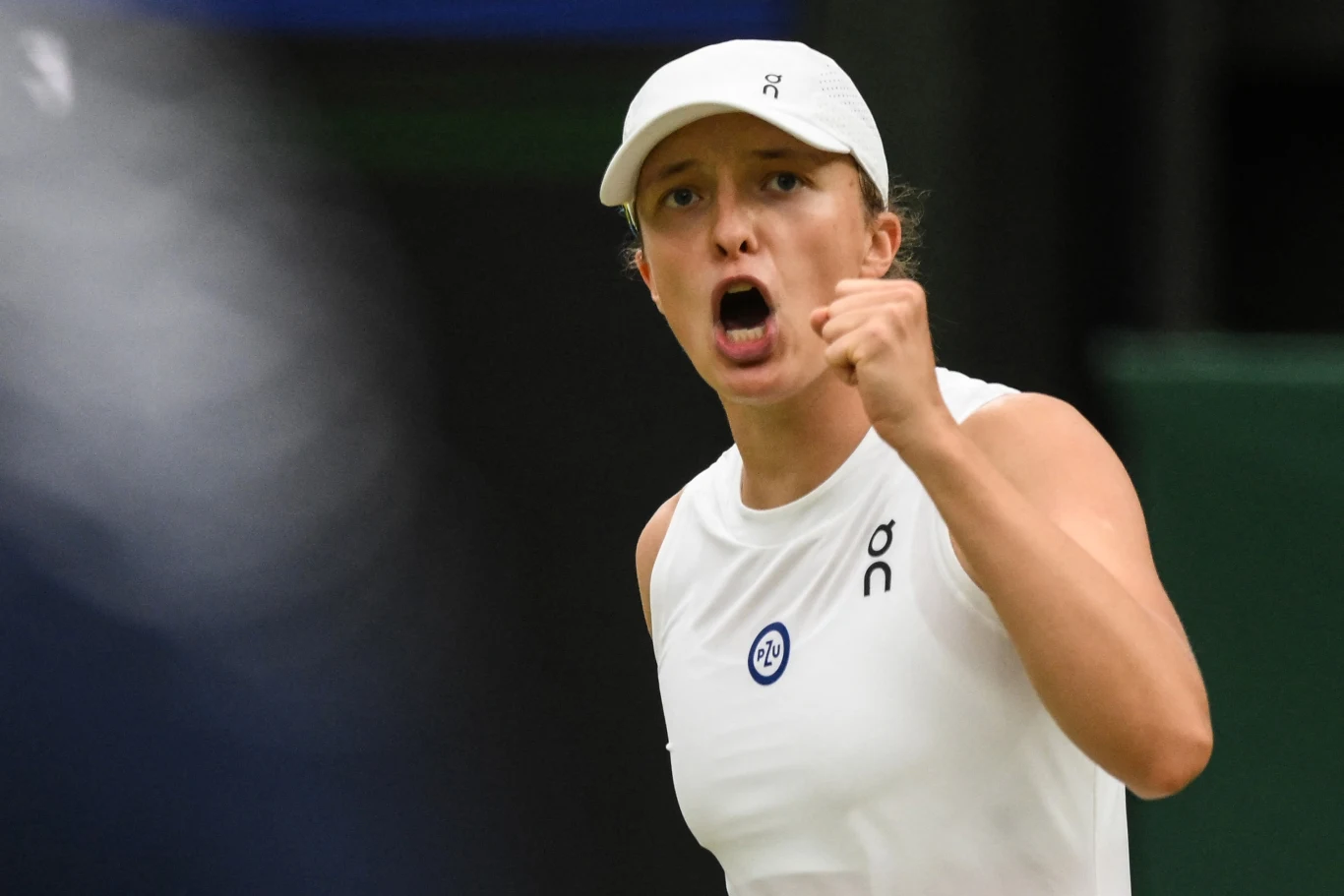 Sportowa zawodniczka w białym stroju i czapce w geście triumfu lub determinacji podczas meczu tenisowego.