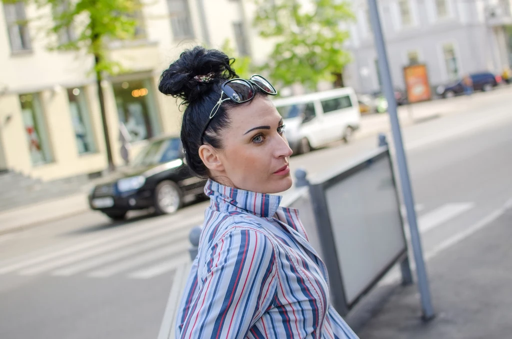 Lata 90 to szczyt popularności imienia Renata Kobieta o ciemnych włosach upiętych w kok oraz przeciwsłonecznych okularach na głowie stoi przy przejściu dla pieszych w centrum miasta, ubrana w koszulę w paski, za nią znajdują się zaparkowane samochody i budynki.