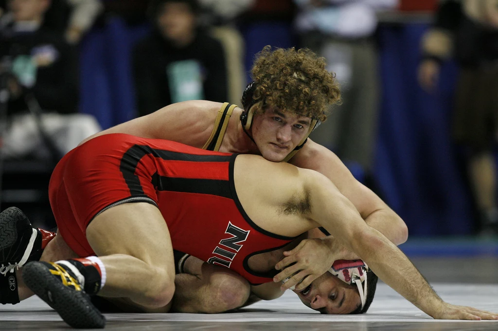 Ben Askren za czasów walk zapaśniczych Dwóch zapaśników podczas walki na macie, zawodnik w czerwonym stroju z napisem 'UNL' znajduje się w pozycji obronnej, natomiast zawodnik w czarno-żółtym stroju kontroluje sytuację, trzymając przeciwnika od góry.