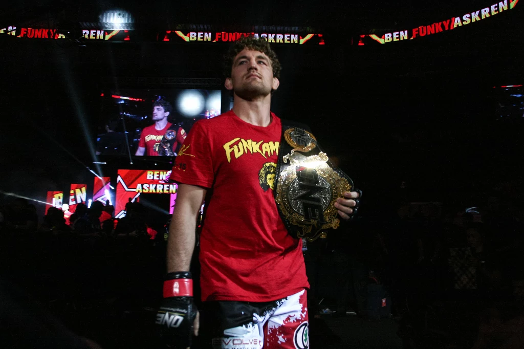 Ben Askren z mistrzowskim pasem ONE Championship Zawodnik MMA ubrany w czerwony t-shirt z napisem Funky oraz spodenki z ochraniaczami stoi na arenie, trzymając pas mistrzowski na ramieniu, w tle widoczny podświetlony ekran z jego wizerunkiem i nazwiskiem.