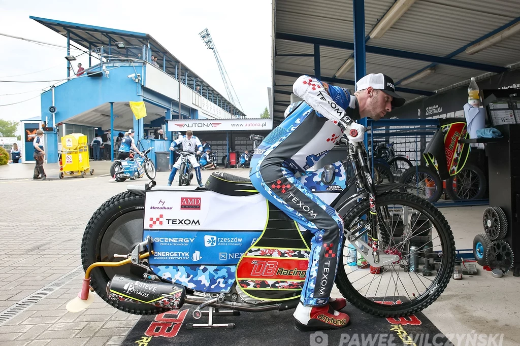 Żużlowiec w sportowym kombinezonie przygotowuje motocykl na parkingu toru żużlowego, w tle widoczne są inne motocykle, akcesoria oraz obsługa techniczna sportowego wydarzenia.