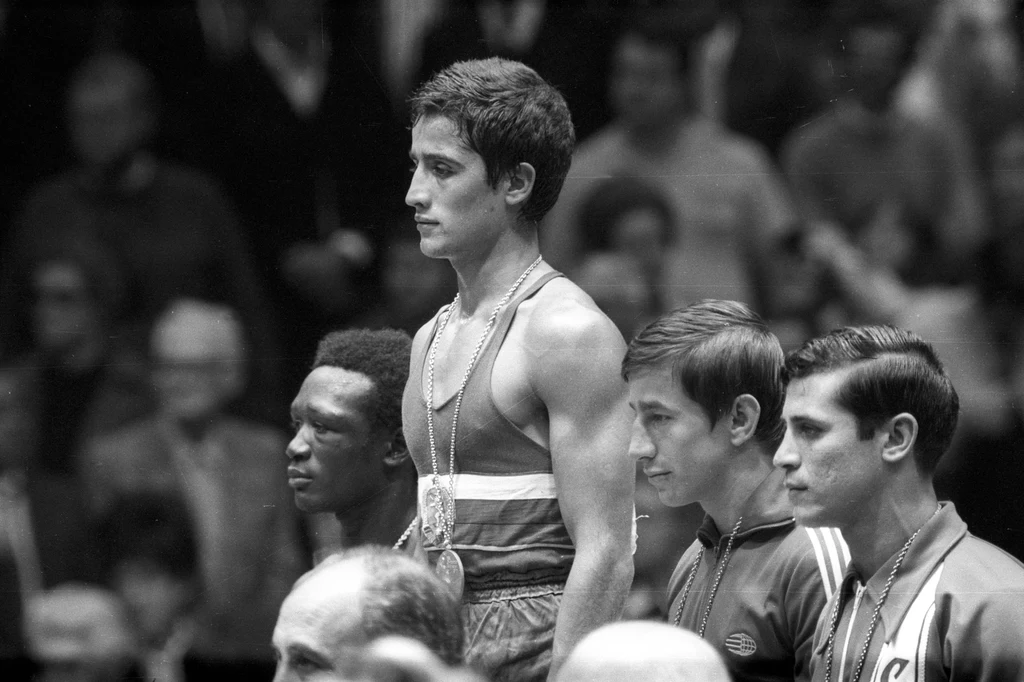 Czterech sportowców na podium podczas ceremonii wręczenia medali, główny zawodnik stoi na najwyższym stopniu, z medalem na szyi, pozostali na niższych stopniach, za nimi widoczna publiczność.