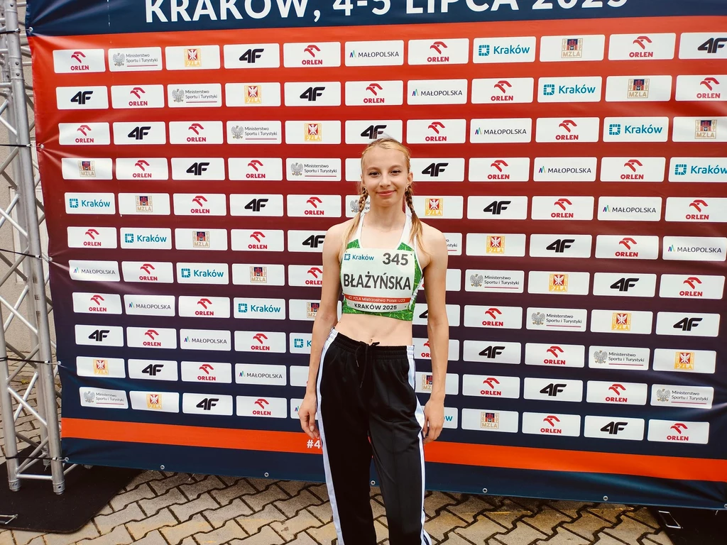 Młoda zawodniczka ubrana w strój sportowy stoi przed ścianką z logotypami sponsorów oraz informacjami o miejscu i dacie wydarzenia sportowego w Krakowie.