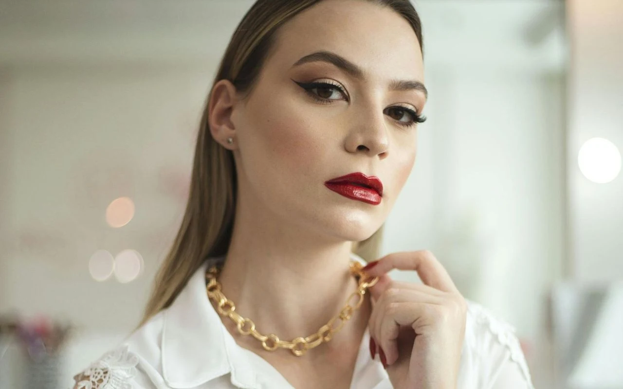 Elegancka kobieta z długimi, prostymi włosami i wyrazistym makijażem, podkreślającym czerwone usta i mocno zarysowane brwi, ubrana w białą koszulę i złoty łańcuszek, patrzy pewnym wzrokiem w stronę obiektywu.