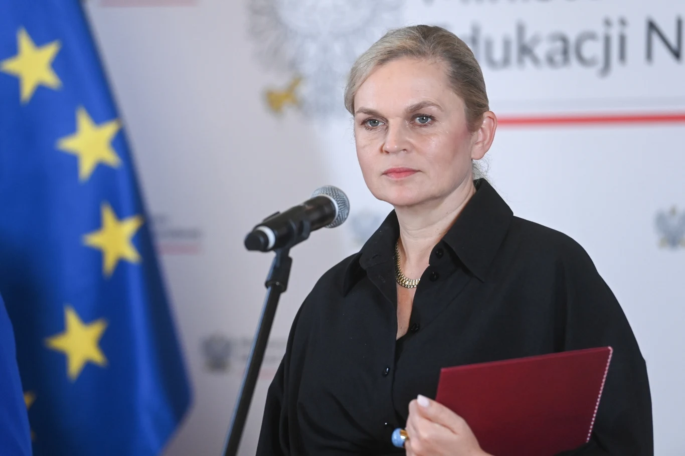 Minister edukacji Barbara Nowacka Kobieta w eleganckiej czarnej koszuli trzyma czerwoną teczkę i stoi przy mikrofonie, w tle widoczna flaga Unii Europejskiej oraz fragment napisu związany z edukacją.