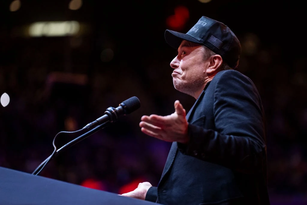 Elon Musk wspierał Donalda Trumpa... a teraz już nie wspiera Mężczyzna w czapce z daszkiem przemawiający do mikrofonu na scenie, gestykulujący rękoma, w tle ciemne, nieostre światła.