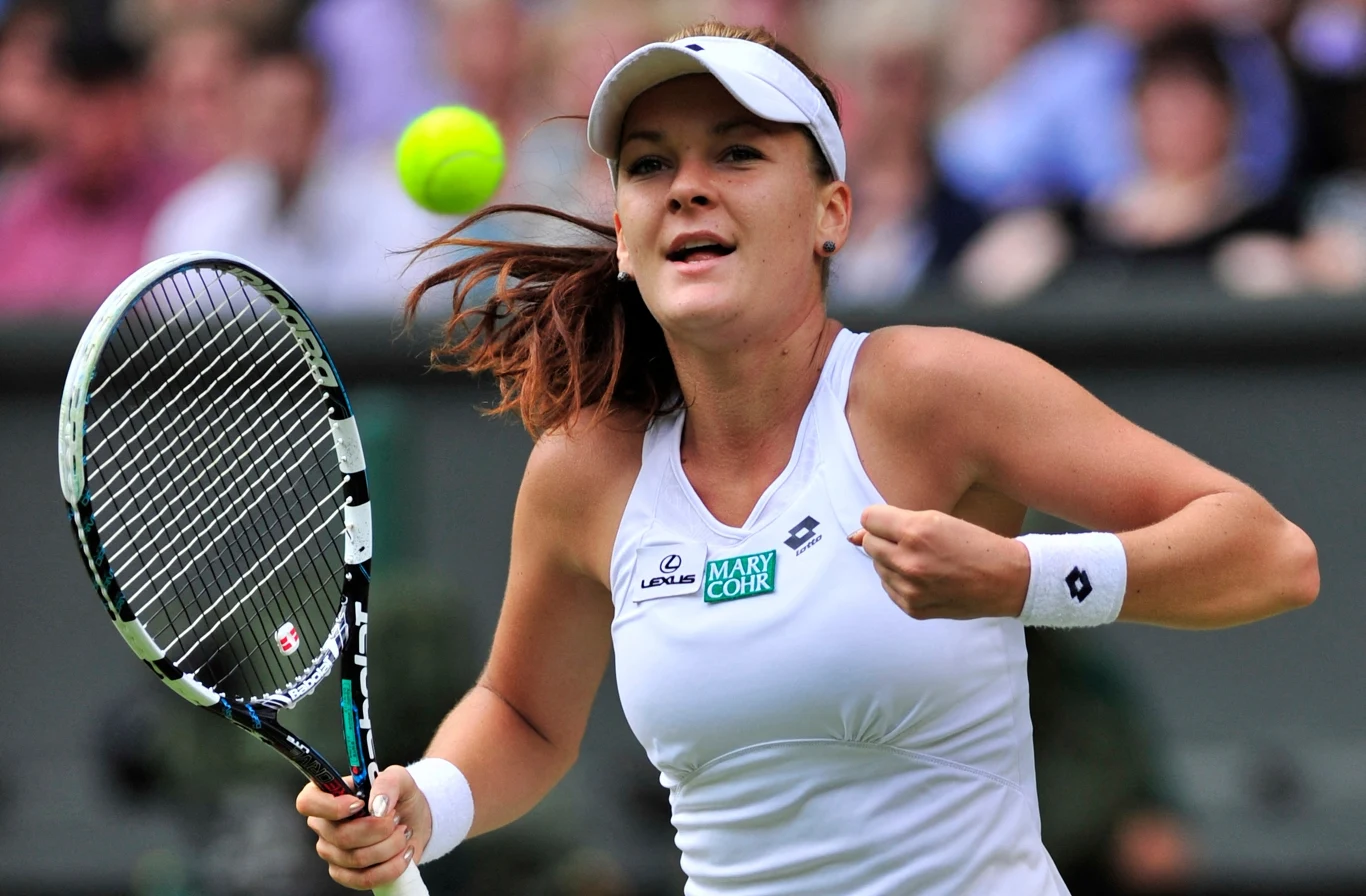 Agnieszka Radwańska po wygranym półfinale Wimbledonu 2012 Tenisistka ubrana w biały strój sportowy koncentruje się na uderzeniu piłki podczas meczu tenisowego, w tle rozmyci widzowie.