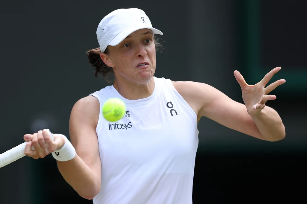 Zawodniczka w białym stroju sportowym podczas intensywnego uderzenia piłki tenisowej na kortach, skoncentrowana i dynamiczna w ruchu, z widoczną piłką tuż przed rakietą.