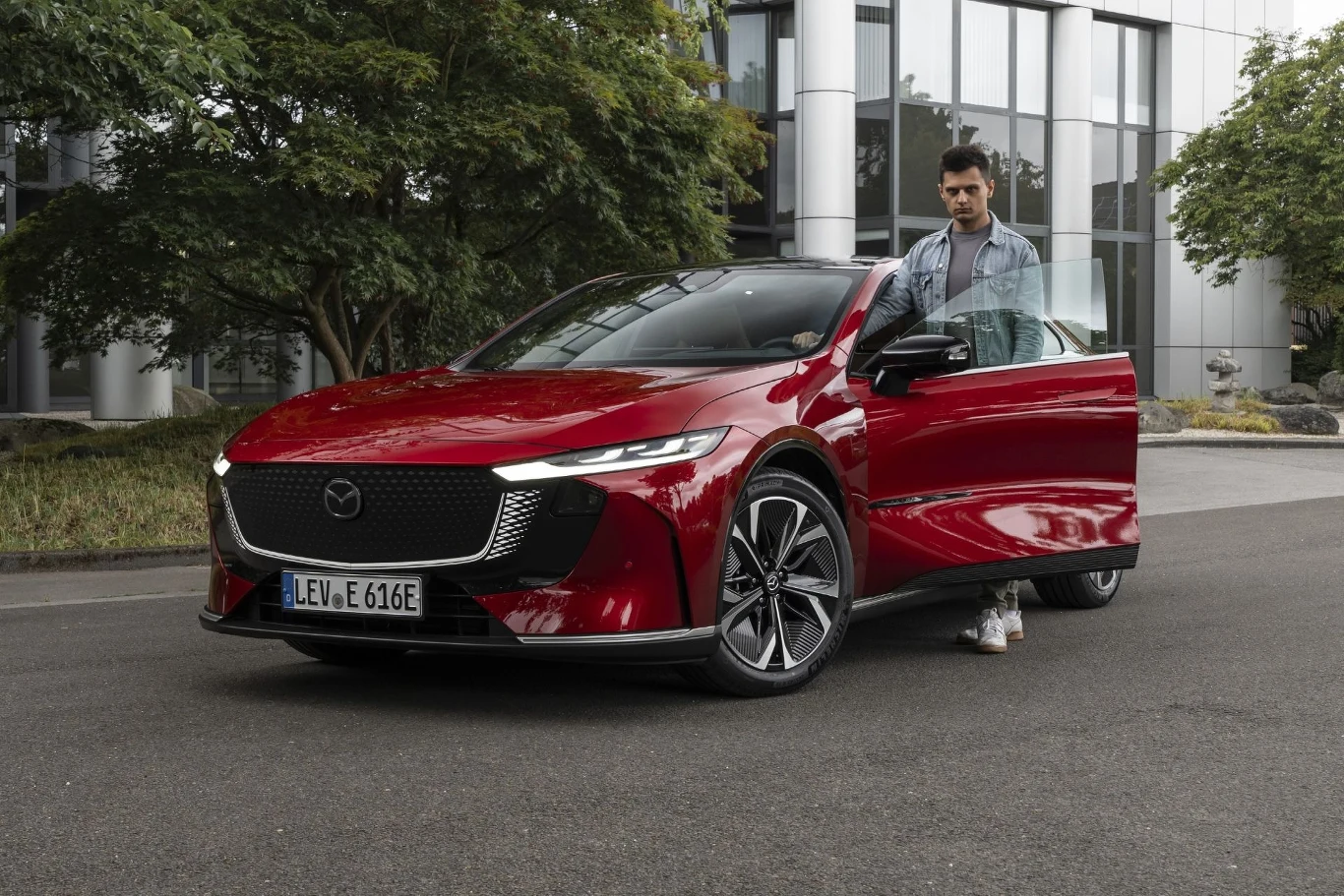 Nowa Mazda 6e to elektryczna interpretacja modelu uwielbianego przez Polaków. Ceną może zamieszać na rynku. Czerwony samochód marki Mazda zaparkowany na ulicy przy nowoczesnym budynku, obok otwartych drzwi samochodu stoi mężczyzna trzymający drzwi, wokół znajduje się zieleń i drzewa.