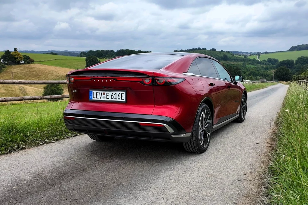 Mazda 6e to de facto chińska Mazda EZ-6. W odróżnieniu od Państwa Środka, w Europie dostępny jest tylko wariant elektryczny. Czerwony samochód elektryczny poruszający się wąską, asfaltową drogą na tle zielonych pól i łagodnych wzgórz, pod pochmurnym niebem.