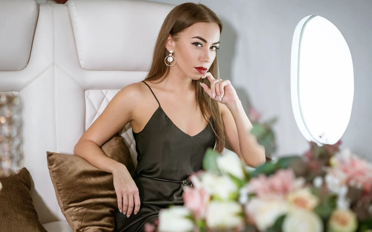 Miłość, kasa i charyzma? Osoby z tymi imionami mają to wszystko Elegancka kobieta w czarnej sukience siedzi na jasnej sofie z poduszką, w tle znajduje się okrągłe oświetlenie oraz nieostre kwiaty na pierwszym planie.