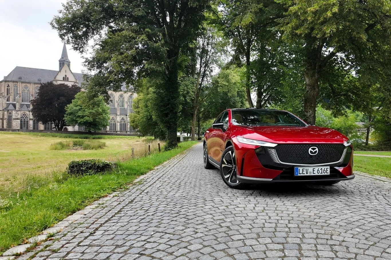 Mazda 6e dostępna jest w Europie w dwóch wersjach napędu - 258 KM i 244 KM. Czerwony samochód marki Mazda na brukowanej drodze otoczonej zielenią, w tle widoczny budynek przypominający kościół z wieżą oraz drzewa po obu stronach ulicy.