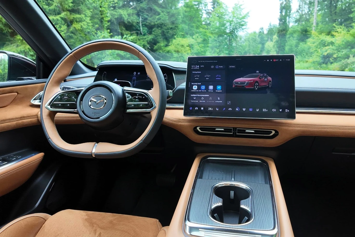 Wnętrze Mazdy 6e utrzymane jest w stylu minimalistycznym - główną rolę odgrywają dwa ekrany: 10,2-calowy zestaw wskaźników i 14,6-calowy wyświetlacz multimediów. Kokpit nowoczesnego samochodu marki Mazda z dużym ekranem dotykowym po prawej stronie, skórzaną kierownicą oraz eleganckim wykończeniem w beżowej kolorystyce, otoczony zielonym krajobrazem widocznym przez szyby.