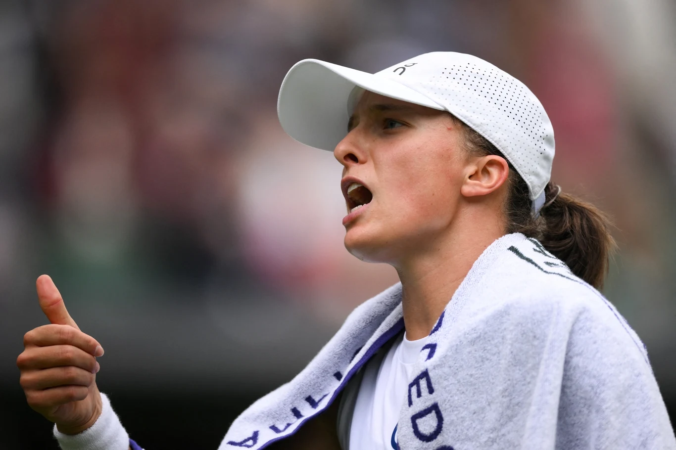 Sportowa emocja, młoda kobieta w białej czapce z daszkiem i ręcznikiem na ramionach energicznie okazuje gest kciuka w górę i wypowiada słowa podczas zawodów sportowych.