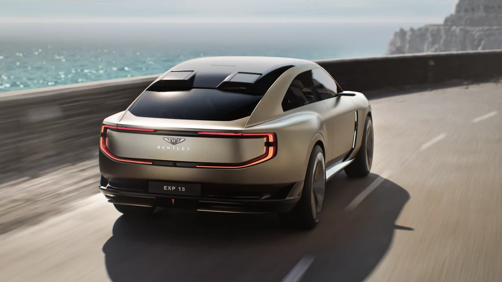 Widok tylnej części nadwozia z nowym znakiem Bentley i światłami w układzie 3D. Nowoczesny samochód koncepcyjny Bentley EXP 15 jedzie dynamicznie wzdłuż nadmorskiej drogi, z lewej strony widać błękitne morze i skalisty brzeg, pojazd prezentuje się elegancko na tle krajobrazu.
