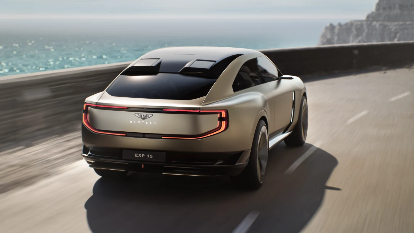 Widok tylnej części nadwozia z nowym znakiem Bentley i światłami w układzie 3D. Nowoczesny samochód koncepcyjny Bentley EXP 15 jedzie dynamicznie wzdłuż nadmorskiej drogi, z lewej strony widać błękitne morze i skalisty brzeg, pojazd prezentuje się elegancko na tle krajobrazu.