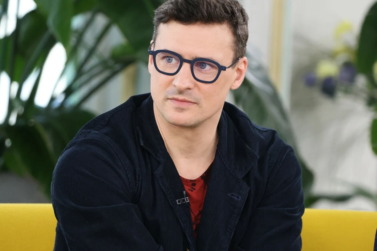 Mateusz Damięcki Mateusz Damięcki w marynarce i okularach siedzi na żółtej kanapie w studiu.