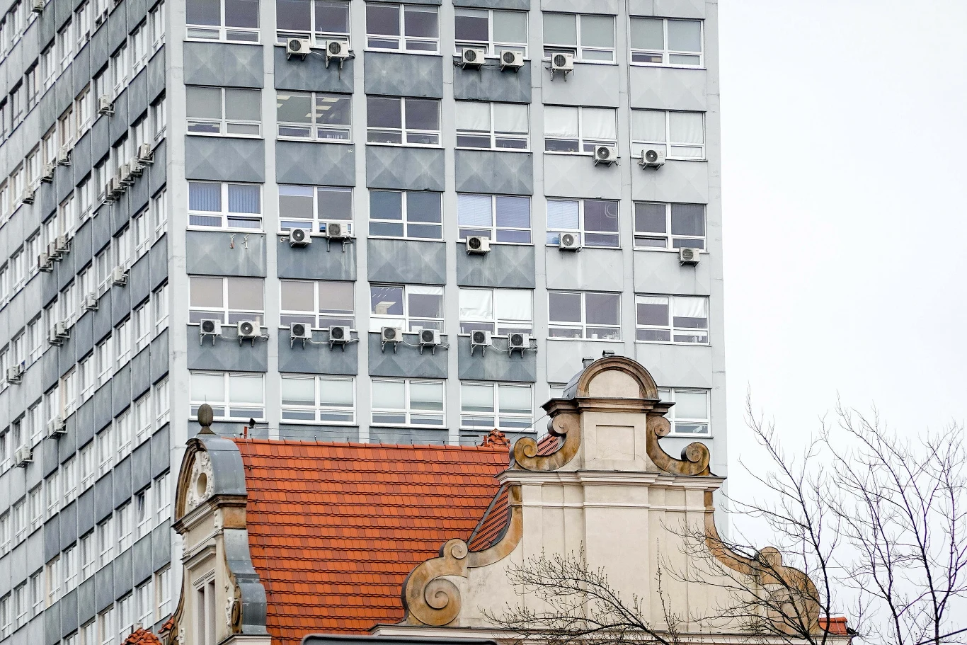 Klimatyzacja może zniszczyć nam życie. Dostrzeżemy to dopiero w przyszłości Kontrast architektoniczny między nowoczesnym, wysokim budynkiem z wieloma klimatyzatorami na fasadzie a ozdobnym, historycznym dachem w stylu klasycznym z czerwonej dachówki.