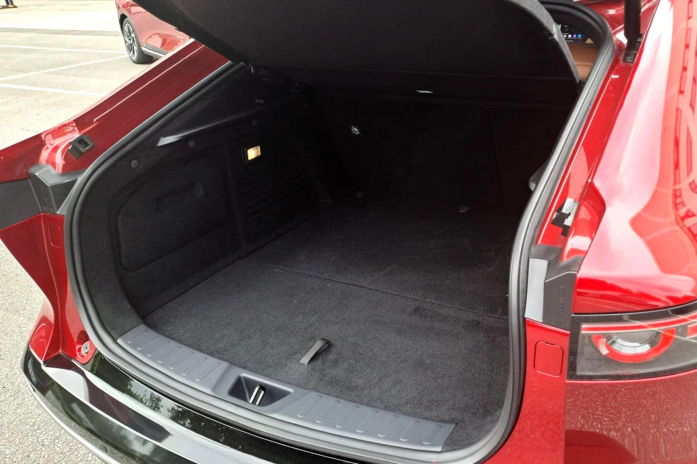 Bagażnik Mazdy 6e oferuje 336 litrów pojemności. Po złożeniu tylnych siedzeń przestrzeń ta rośnie do 678 litrów. Otwarty bagażnik samochodu osobowego o czerwonym kolorze nadwozia, wykończony czystą, czarną tapicerką i widoczny fragment tylnego światła.