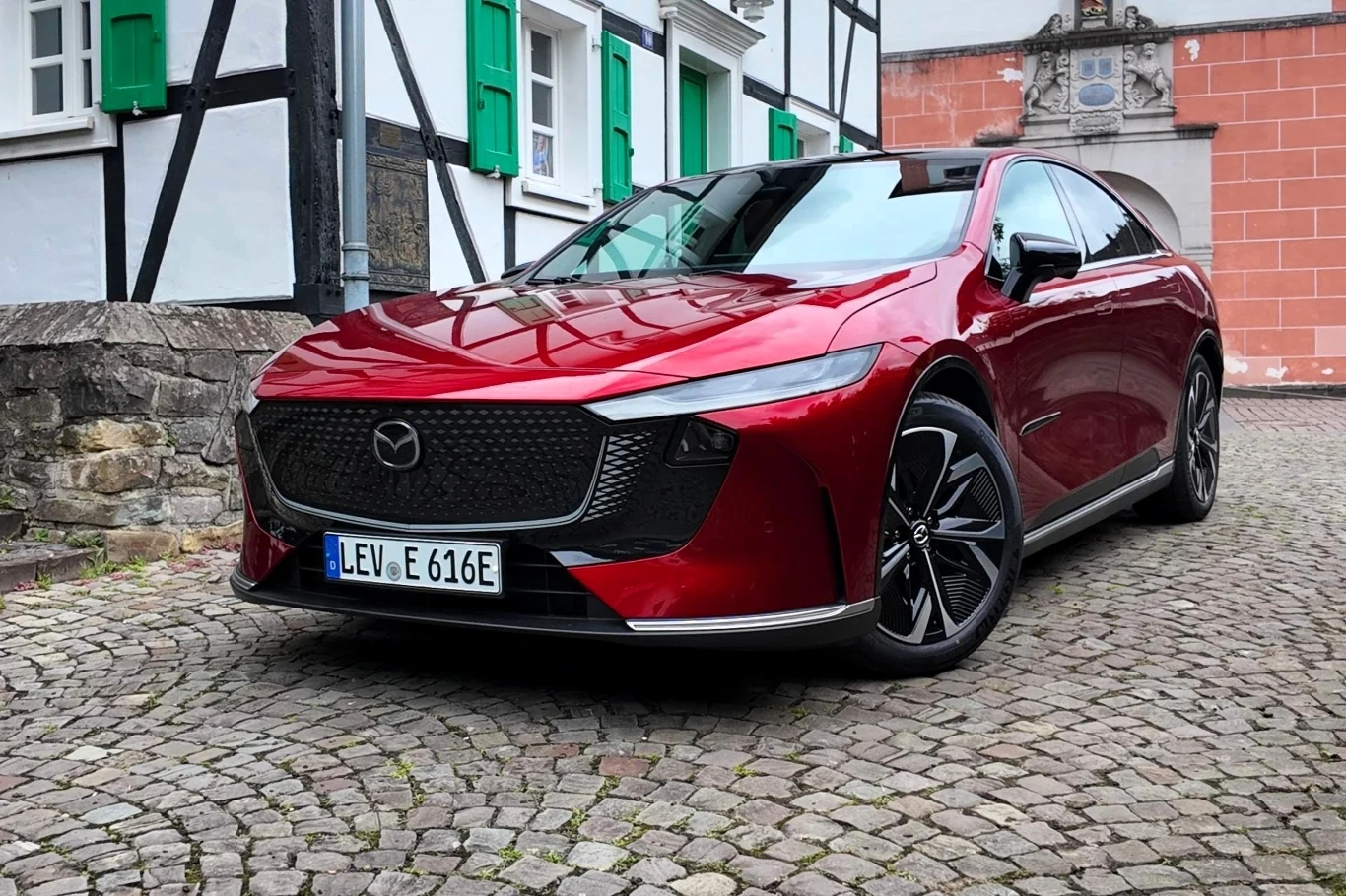 Dzięki akumulatorowi ukrytemu w podłodze oraz rozkładowi masy przód:tył na poziomie 47:53, Mazda 6e jest bardzo stabilna w zakrętach. Czerwony nowoczesny samochód marki Mazda stoi zaparkowany na brukowanej uliczce, w tle widoczne zabytkowe budynki z białymi ścianami, czarnymi belkami i zielonymi okiennicami.