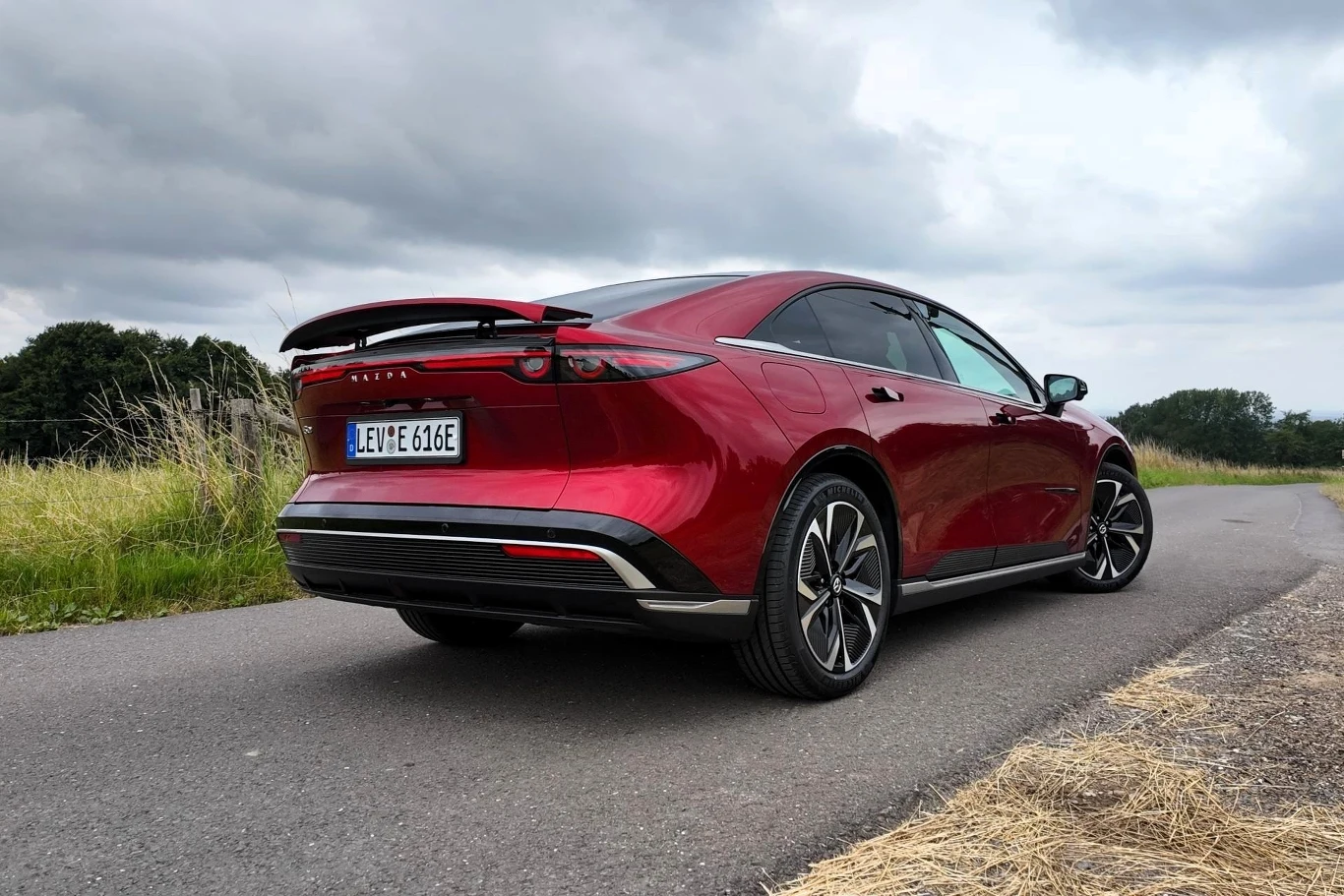 Mazda 6e wyposażona jest w spoiler, który wysuwa się przy prędkości 90 km/h i chowa przy prędkości 50 km/h. Możemy nim jednak sterować także "ręcznie". Czerwony samochód osobowy typu sedan z nowoczesnym designem, stojący na asfaltowej drodze w otoczeniu zieleni i pola, pod pochmurnym niebem.