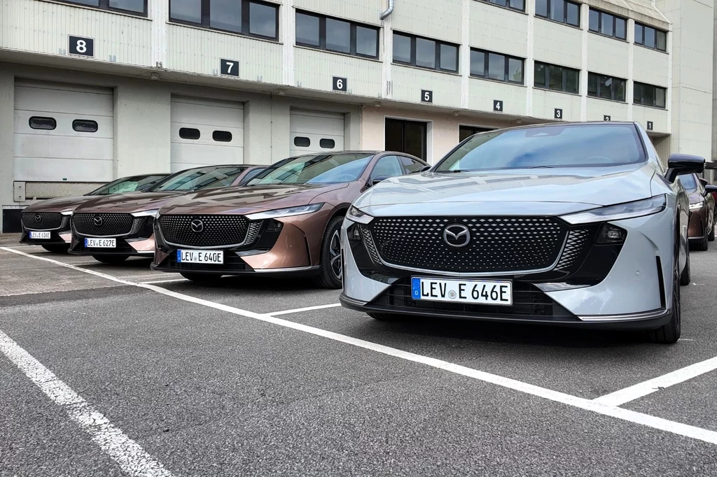 Nowa Mazda 6e startuje w Polsce od 198 200 zł. Nowoczesne samochody marki Mazda stojące równolegle na parkingu przed budynkiem przemysłowym, z widocznymi numerami na bramach garażowych i dużymi oknami w tle.