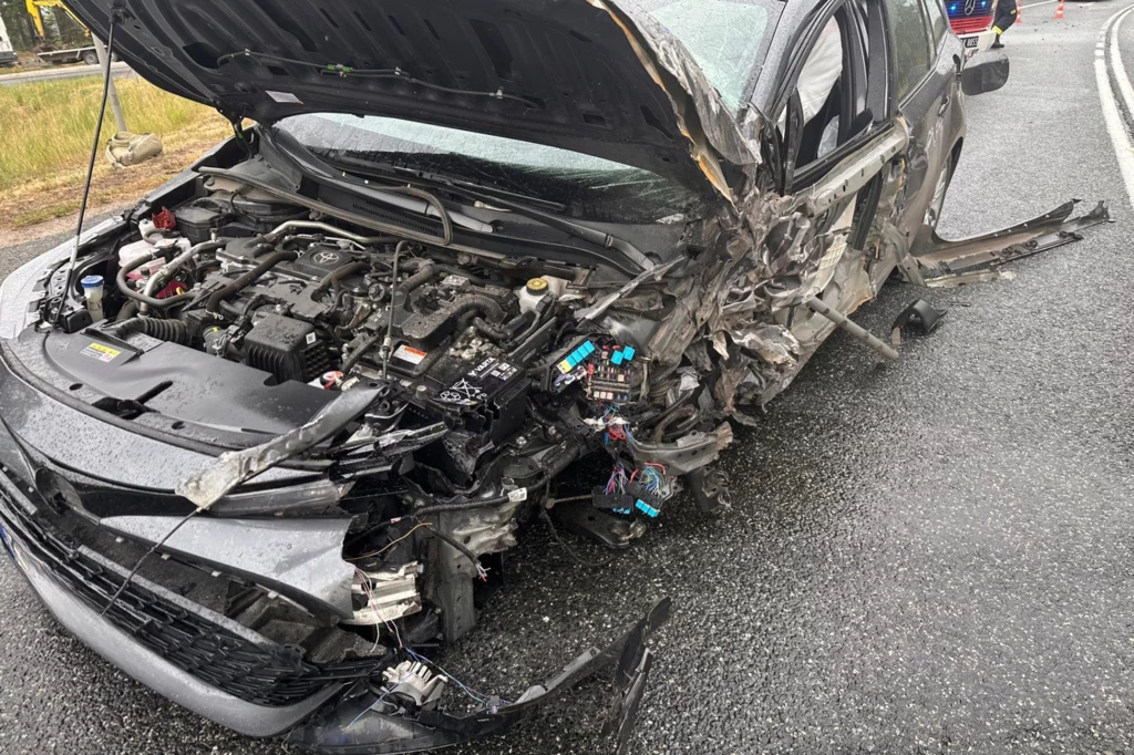 Kierowcy Toyoty na szczęście nic się nie stało. Rozbity samochód osobowy z poważnie uszkodzonym przodem i otwartą maską, widoczne części silnika oraz uszkodzone elementy karoserii na mokrej nawierzchni drogi, w tle fragment samochodu służb ratunkowych.