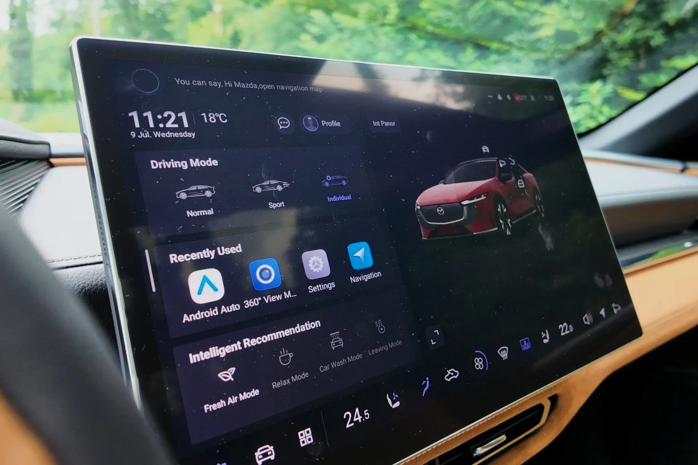 Zdecydowaną liczbą funkcji w Maździe 6e, w tym światłami czy trybami jazdy, sterujemy z poziomu 14,6-calowego ekranu multimediów. Duży ekran dotykowy samochodowego systemu multimedialnego wyświetlający różne tryby jazdy, aktualną godzinę i temperaturę, menu z poleceniami głosowymi, skrótami do Android Auto, nawigacji oraz ustawieniami pojazdu, z widoczną grafiką pojazdu po prawej...