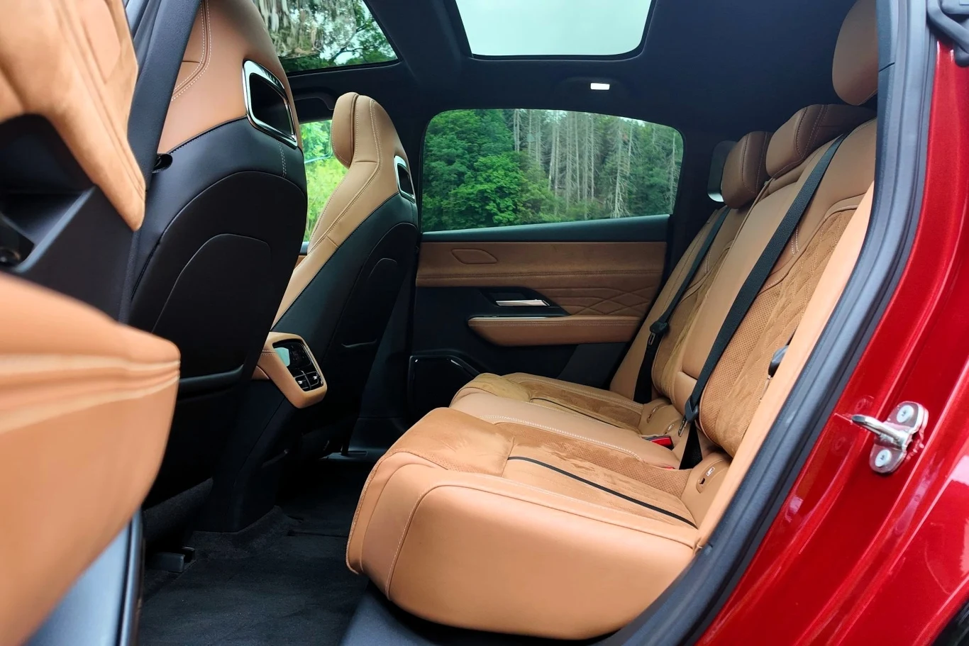 Mazda 6e ma 4 921 mm długości i rozstaw osi na poziomie 2 895 mm. Przekłada się to wymiernie na przestrzeń w drugim rzędzie siedzeń. Miejsca jest pod dostatkiem. Luksusowe, przestronne wnętrze samochodu z jasnobrązowymi skórzanymi siedzeniami, panoramiczny dach szklany, zielony las widoczny przez okno, eleganckie wykończenia drzwi i tylnej części foteli.