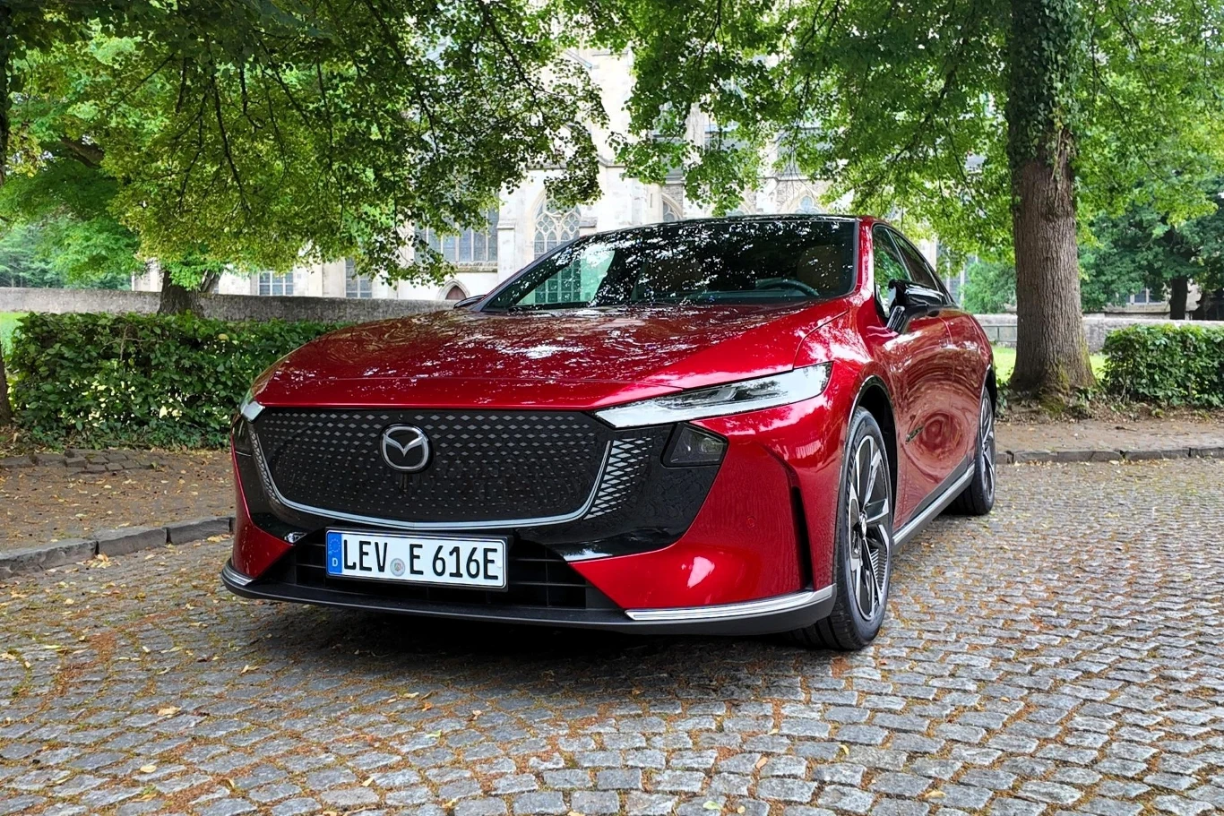 Mazda 6e dostępna jest tylko z napędem elektrycznym. Są dwie opcje akumulatora: 68,8 kWh oraz 80 kWh. W wersji z większą baterią auto przejedzie do 552 km w cyklu mieszanym. Czerwony samochód marki Mazda stoi na brukowanej drodze pod dużym drzewem, otoczony zielenią i niewysokim murem z kamieni, widoczne są wyraźnie reflektory oraz czarna osłona chłodnicy z logo Mazdy.